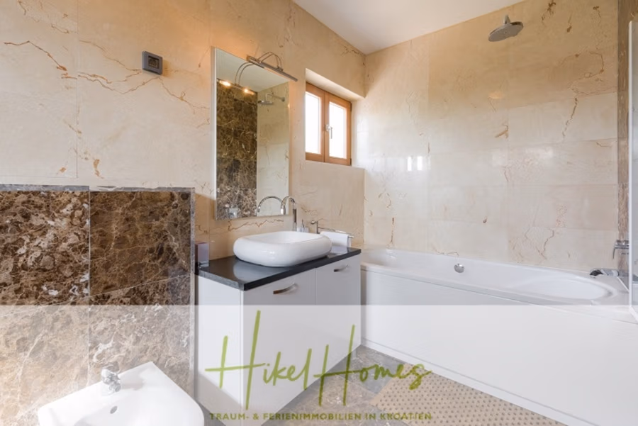 Villa mit 3 großzügigen Appartements mit jeweils 3 SZ, Küche, 2 x Pool, … - Modernes Badezimmer mit beigen Marmorwänden und einem quadratischen Fenster. Es verfügt über ein weißes Waschbecken mit großem Spiegel, eine Badewanne und ein Bidet. Die graue Waschtischplatte und die teilweise sichtbare Dusche stehen vor einer dunklen Marmorwand. Logotext am unteren Rand. (41145)