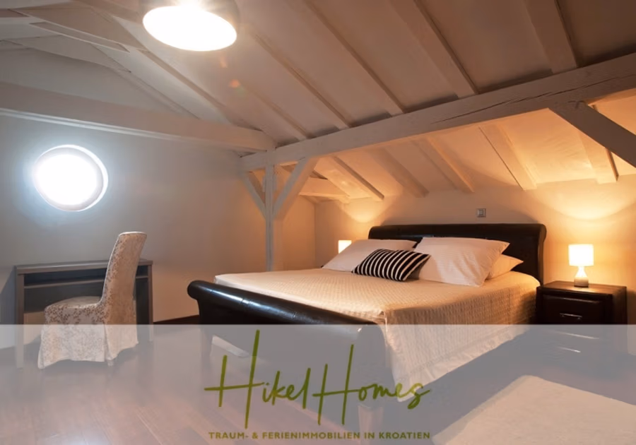 Villa mit 3 großzügigen Appartements mit jeweils 3 SZ, Küche, 2 x Pool, … - Dachgeschoss-Schlafzimmer mit schräger Decke und Holzbalken, ausgestattet mit einem Doppelbett mit Kissen und einem Nachttisch mit Lampe. Ein kleiner Schreibtisch mit Stuhl steht neben einem runden Fenster. Der Text lautet „Hikel Homes – Traum- & Ferienimmobilien in Kroatien“. (41148)