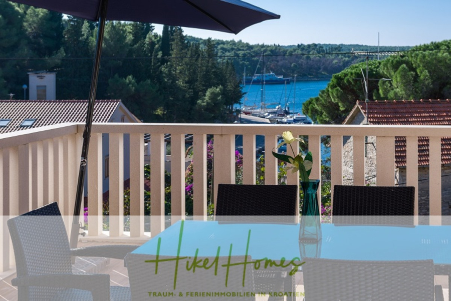 Villa mit 3 großzügigen Appartements mit jeweils 3 SZ, Küche, 2 x Pool, … - Eine Terrasse mit einem Glastisch, schwarzen Stühlen und einer Vase blickt auf einen Yachthafen mit Booten, umgeben von Bäumen und Dächern, unter einem blauen Sonnenschirm. Im Hintergrund sind das Meer und bewaldete Hügel zu sehen. (46274)