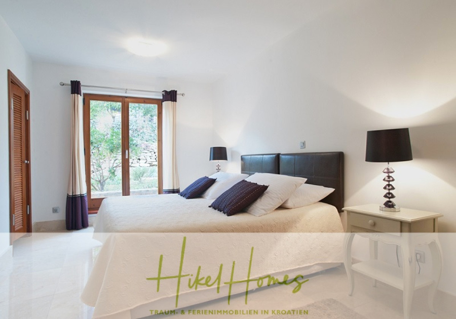 Villa mit 3 großzügigen Appartements mit jeweils 3 SZ, Küche, 2 x Pool, … - Helles Schlafzimmer mit einem großen Bett, weißer Bettwäsche, zwei Nachttischen mit schwarzen Lampen, Vorhängen, die eine Glastür zu einem Garten einrahmen, und dem Logo "Hikel Homes" mit deutschem Text am unteren Rand. (46273)