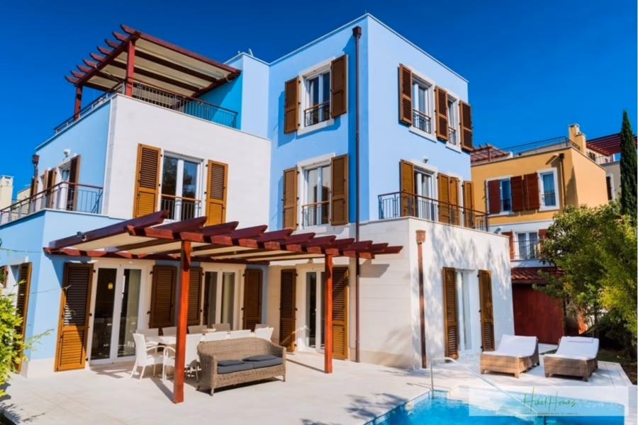 Großzügige Villa im Mittelmeerstil mit 3 SZ, Pool, Dachterrasse mit Meerblick (12) - Ein modernes zweistöckiges Haus mit hellblauen Wänden, hölzernen Fensterläden und mehreren Balkonen. Eine Pergola überdacht eine Terrasse mit Gartenmöbeln. Im Vordergrund befindet sich ein kleiner Pool vor dem Hintergrund des klaren blauen Himmels. (42155)