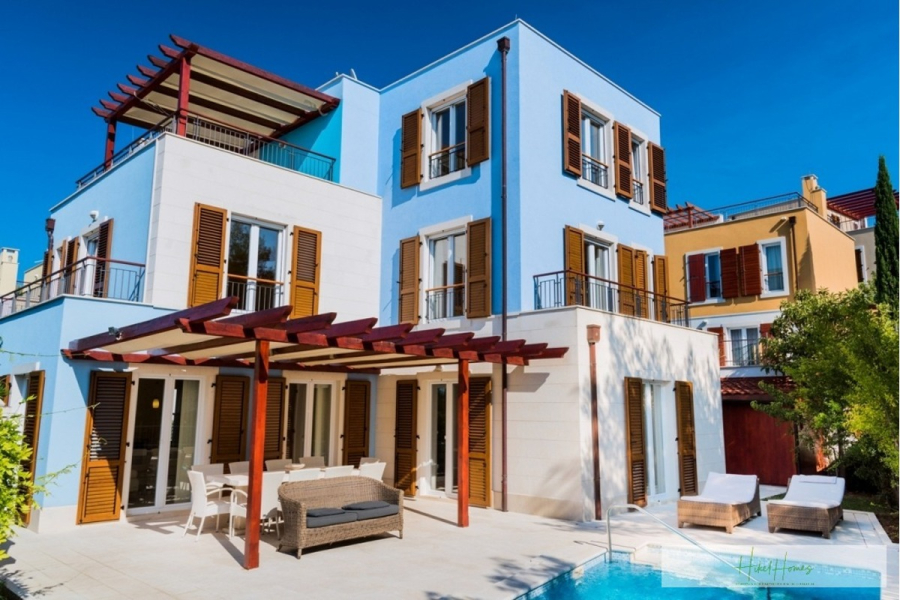 Großzügige Villa im Mittelmeerstil mit 3 SZ, Pool, Dachterrasse mit Meerblick (12) - Ein modernes, dreistöckiges, blau-weißes Haus mit hölzernen Fensterläden, mehreren Balkonen, einer Pergola-überdachten Terrasse mit Gartenmöbeln und einem Swimmingpool unter einem strahlend blauen Himmel. (45881)