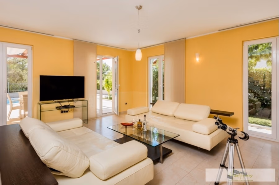 Großzügige Villa im Mittelmeerstil mit 3 SZ, Pool, Dachterrasse mit Meerblick (12) - Ein modernes Wohnzimmer mit gelben Wänden, cremefarbenen Ledersofas und einem Couchtisch aus Glas. Ein Flachbildfernseher steht auf einem Glasständer. Große Fenster geben den Blick auf die grüne Umgebung frei, und neben den Glastüren zur Terrasse steht ein Teleskop. (42161)