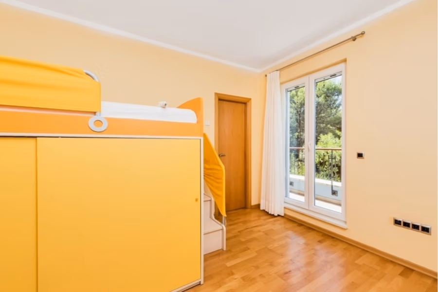 Großzügige Villa im Mittelmeerstil mit 3 SZ, Pool, Dachterrasse mit Meerblick (12) - Ein heller, minimalistischer Raum mit Holzboden, einem gelben Etagenbett mit eingebautem Stauraum und einer Treppe. Eine Glastür mit weißen Vorhängen öffnet sich zu einem Balkon mit Blick ins Grüne. Die Wände sind hellgelb gestrichen. (42175)
