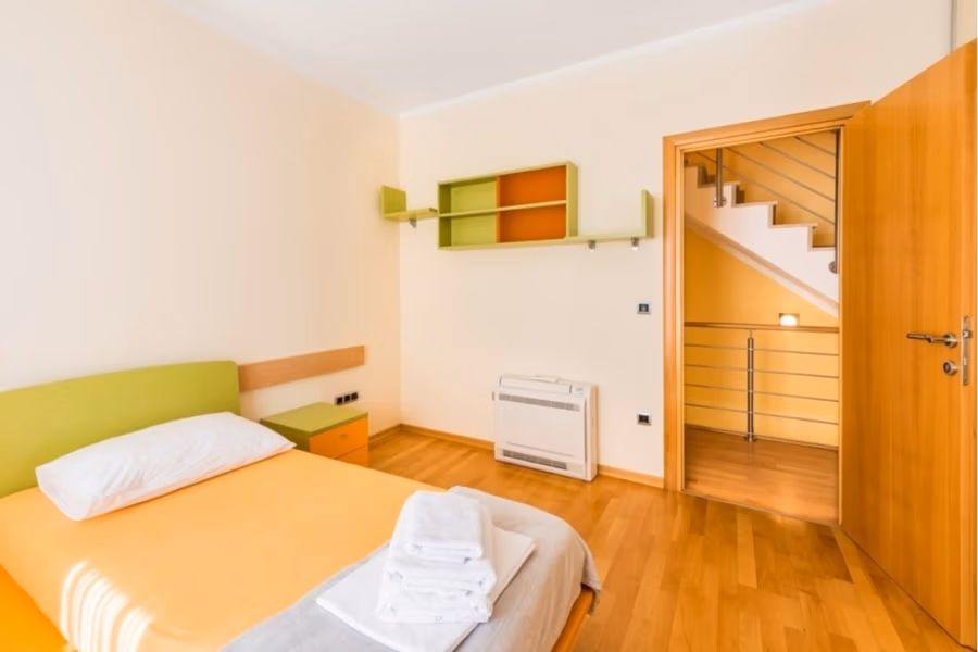 Großzügige Villa im Mittelmeerstil mit 3 SZ, Pool, Dachterrasse mit Meerblick (12) - Ein kleines Schlafzimmer mit einem Einzelbett mit orangefarbener Bettwäsche und grünem Kopfteil. Neben dem Bett steht ein Nachttisch mit gefalteten Handtüchern. Die Wände sind beige mit grünen und orangefarbenen Regalen. Eine offene Tür gibt den Blick auf eine Treppe mit Metallgeländer frei. (42173)