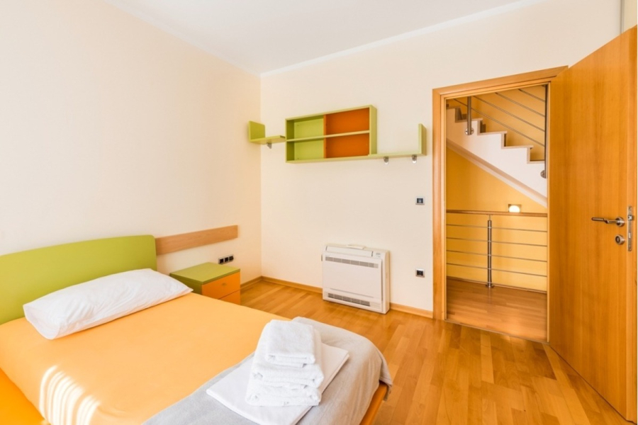 Großzügige Villa im Mittelmeerstil mit 3 SZ, Pool, Dachterrasse mit Meerblick (12) - Ein modernes, minimalistisches Schlafzimmer mit einem Einzelbett mit orangefarbener Bettwäsche, einem gefalteten Handtuch darauf, hellem Holzboden, grünen und orangefarbenen Möbelakzenten und einer offenen Tür, die eine Treppe zeigt. (45899)