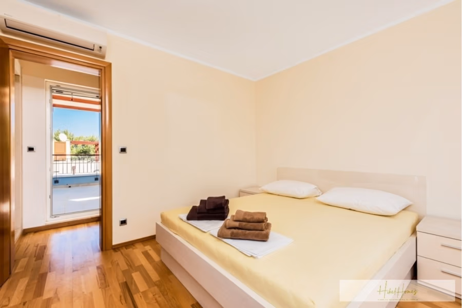 Großzügige Villa im Mittelmeerstil mit 3 SZ, Pool, Dachterrasse mit Meerblick (12) - Ein minimalistisches Schlafzimmer mit einem Doppelbett, bedeckt mit einem hellgelben Laken, zwei weißen Kissen und gefalteten Handtüchern am Fußende. Heller Holzboden, eine Tür führt auf einen Balkon mit teilweisem Blick ins Grüne. (42168)
