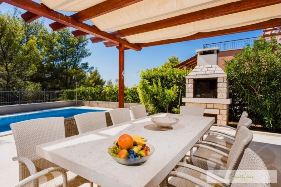 Großzügige Villa im Mittelmeerstil mit 3 SZ, Pool, Dachterrasse mit Meerblick (12) - Außenterrasse mit großem Esstisch. Eine Schale mit frischem Obst steht darauf. Der Bereich wird von einer Pergola beschattet, und ein eingebauter Steingrill befindet sich in der Nähe. Im Hintergrund sind üppiges Grün und ein Swimmingpool zu sehen. (42167)