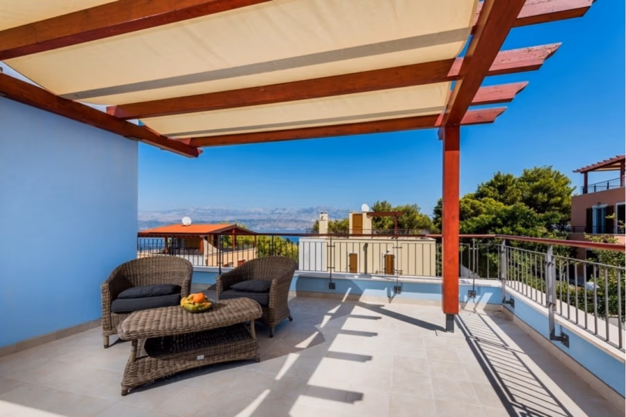 Großzügige Villa im Mittelmeerstil mit 3 SZ, Pool, Dachterrasse mit Meerblick (12) - Eine Dachterrasse mit Korbmöbeln und einem kleinen Tisch unter einer hölzernen Pergola. Von der Terrasse aus hat man einen malerischen Blick auf das Meer und die umliegenden Gebäude, umgeben von Bäumen. Der Himmel ist klar und blau. (42178)