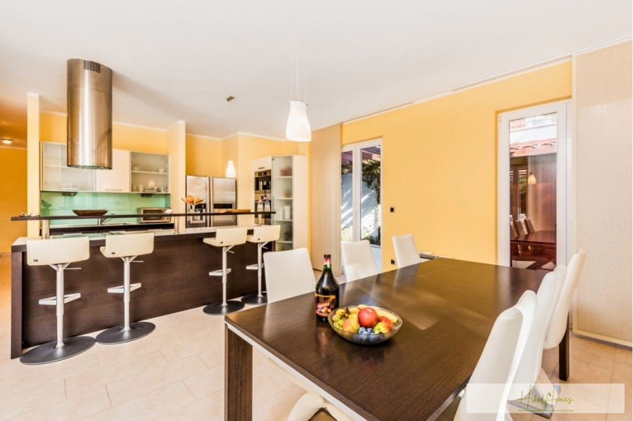 Großzügige Villa im Mittelmeerstil mit 3 SZ, Pool, Dachterrasse mit Meerblick (12) - Moderne Küche und Essbereich mit gelben Wänden, einem Esstisch aus dunklem Holz mit weißen Stühlen, einer Obstschale und einer Flasche. Im Hintergrund ist eine Kücheninsel mit Barhockern und Geräten aus Edelstahl zu sehen. (45882)