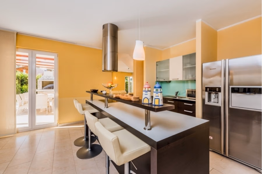 Großzügige Villa im Mittelmeerstil mit 3 SZ, Pool, Dachterrasse mit Meerblick (12) - Moderne Küche mit gelben Wänden, einer Kücheninsel mit weißen Barhockern, Edelstahlgeräten und grüner Küchenrückwand. Eine Glastür führt auf eine Terrasse mit rot-weiß gestreifter Markise. Über der Kücheninsel hängen Pendelleuchten. (42181)
