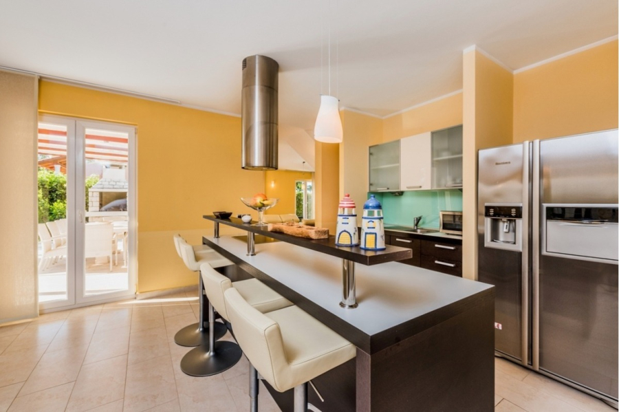 Großzügige Villa im Mittelmeerstil mit 3 SZ, Pool, Dachterrasse mit Meerblick (12) - Moderne Küche mit gelben Wänden, einer langen Insel mit beigen Barhockern, Edelstahlgeräten, Hängeleuchten und großen Glastüren, die zu einer Terrasse im Freien führen, auf der Grünflächen zu sehen sind. (45907)