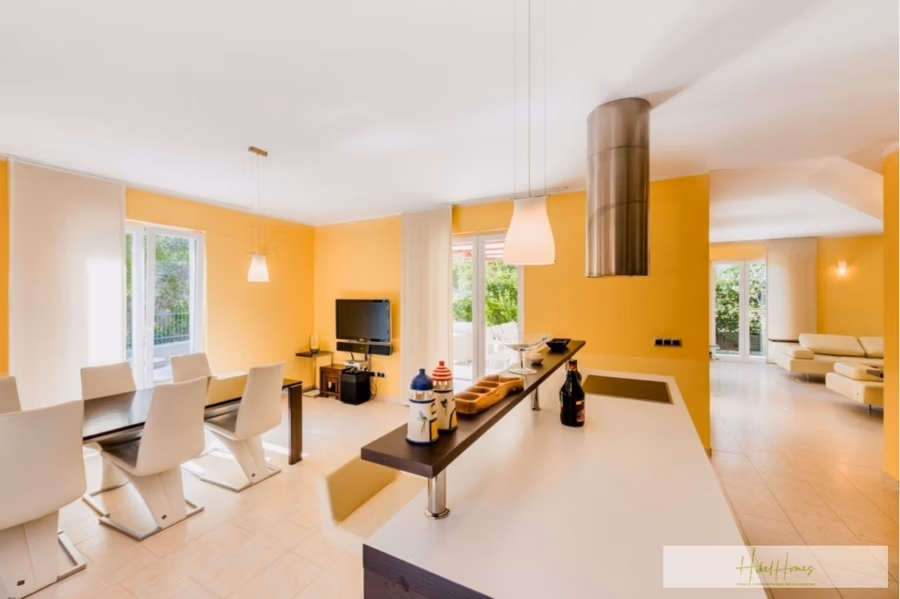 Großzügige Villa im Mittelmeerstil mit 3 SZ, Pool, Dachterrasse mit Meerblick (12) - Großzügiges, modernes Wohnzimmer und Küche mit gelben Wänden und weißen Bodenfliesen. Es verfügt über einen Esstisch mit weißen Stühlen, einen Fernseher und eine Kücheninsel mit Barhockern. Große Fenster und Schiebetüren sorgen für Tageslicht. (42163)