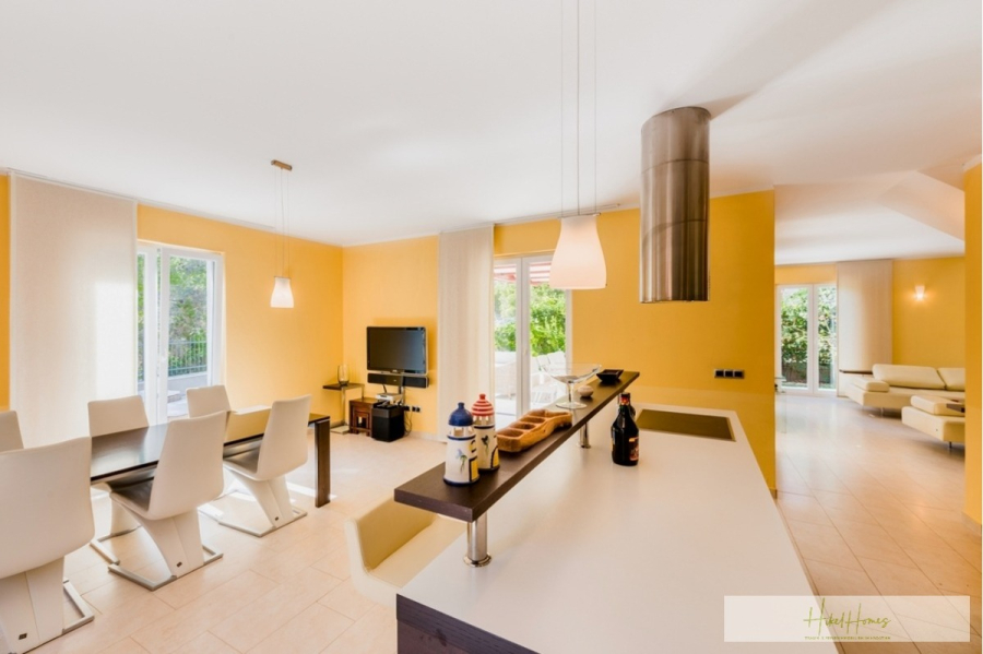 Großzügige Villa im Mittelmeerstil mit 3 SZ, Pool, Dachterrasse mit Meerblick (12) - Ein heller, moderner, offener Wohnbereich mit gelben Wänden, einem Essbereich mit weißen Stühlen, einer Kücheninsel mit Hängeleuchten und einer gemütlichen Sitzecke mit großen Fenstern und Glastüren nach draußen. (45889)