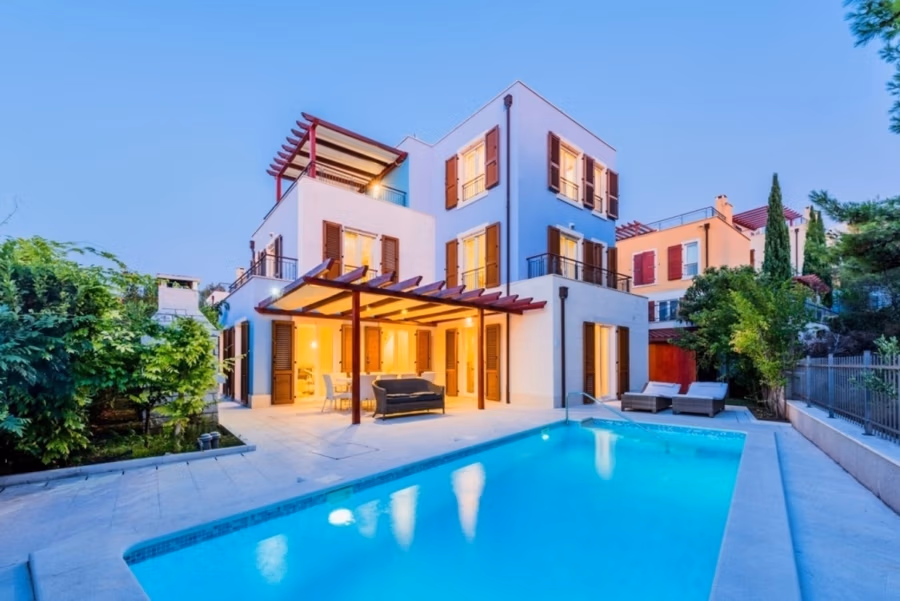 Großzügige Villa im Mittelmeerstil mit 3 SZ, Pool, Dachterrasse mit Meerblick (12) - Ein modernes, dreistöckiges Haus mit großen Fenstern und Balkon wird in der Dämmerung beleuchtet. Die großzügige Terrasse bietet Sitzgelegenheiten unter einer Pergola. Im Vordergrund befindet sich ein leuchtend blauer Pool, umgeben von üppigem Grün. (42172)