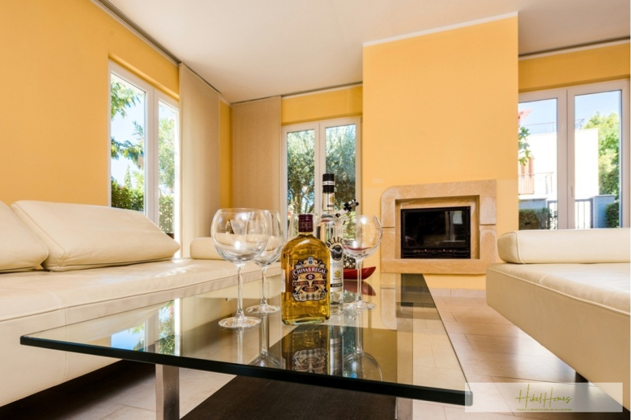 Großzügige Villa im Mittelmeerstil mit 3 SZ, Pool, Dachterrasse mit Meerblick (12) - Ein helles Wohnzimmer mit gelben Wänden, cremefarbenen Sofas, einem gläsernen Couchtisch, auf dem eine Schnapsflasche und Gläser stehen, und einem Kamin. Große Fenster lassen das Sonnenlicht herein und geben den Blick auf das Grün draußen frei. (45886)
