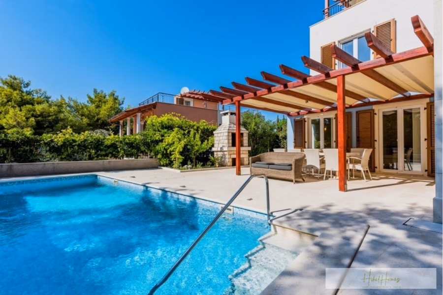 Großzügige Villa im Mittelmeerstil mit 3 SZ, Pool, Dachterrasse mit Meerblick (12) - Ein luxuriöser Außenbereich mit rechteckigem Swimmingpool, einer Terrasse mit roter Pergola, Loungemöbeln aus Korbgeflecht und einem Esstisch mit Stühlen. Umgeben von viel Grün und strahlend blauem Himmel wirkt die Szene hell und einladend. (42158)