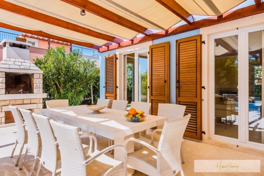 Großzügige Villa im Mittelmeerstil mit 3 SZ, Pool, Dachterrasse mit Meerblick (12) - Außenterrasse mit weißem Esstisch und Korbstühlen unter einer Holzpergola. Der Tisch ist mit einer Obstschale gedeckt. In der Nähe ist ein gemauerter Grill zu sehen. Der Bereich ist von Grün umgeben und führt durch Glastüren in den Innenbereich. (42165)