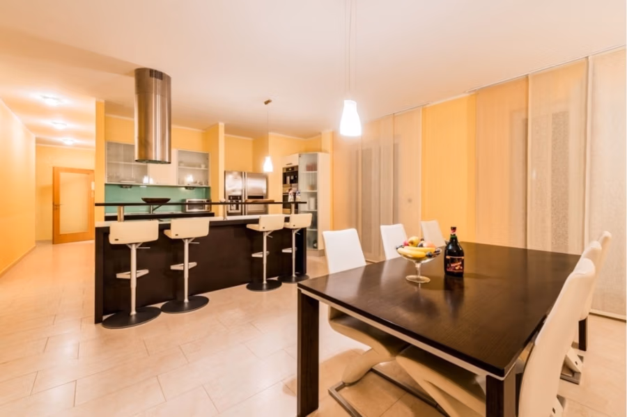 Großzügige Villa im Mittelmeerstil mit 3 SZ, Pool, Dachterrasse mit Meerblick (12) - Moderne Küche mit langem Esstisch aus Holz und weißen Stühlen. Der Raum verfügt über eine elegante schwarze Kücheninsel mit Barhockern, Deckenbeleuchtung und beigefarbenen Wänden. Auf dem Tisch stehen eine Obstschale und eine Flasche, an der Decke befindet sich eine Einbauleuchte. (42171)