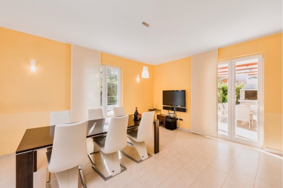 Großzügige Villa im Mittelmeerstil mit 3 SZ, Pool, Dachterrasse mit Meerblick (12) - Ein helles Esszimmer mit hellorangen Wänden ist mit einem dunklen Holztisch und sechs weißen Stühlen ausgestattet. An der rechten Wand hängt ein Flachbildfernseher. Sonnenlicht fällt durch Fenster und Glastüren ein und beleuchtet den gefliesten Boden und die Terrasse. (42182)