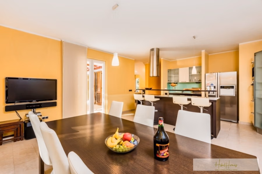 Großzügige Villa im Mittelmeerstil mit 3 SZ, Pool, Dachterrasse mit Meerblick (12) - Moderne Küche und Essbereich mit einem Holztisch, auf dem eine Obstschale und eine Flasche stehen. Weiße Stühle umgeben den Tisch. Die Küche verfügt über eine elegante Kochinsel, Edelstahlgeräte und leuchtend gelbe Wände. Im Hintergrund ist ein wandmontierter Fernseher zu sehen. (42157)