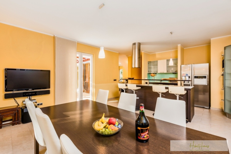 Großzügige Villa im Mittelmeerstil mit 3 SZ, Pool, Dachterrasse mit Meerblick (12) - Moderner offener Küchen- und Essbereich mit gelben Wänden, einem dunklen Holztisch für sechs Personen, einer Schale mit Obst und einer Flasche auf dem Tisch, Barhockern am Tresen und Geräten aus Edelstahl. (45883)