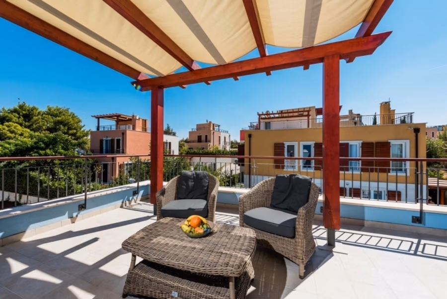 Großzügige Villa im Mittelmeerstil mit 3 SZ, Pool, Dachterrasse mit Meerblick (12) - Eine Dachterrasse mit Korbmöbeln, darunter zwei Stühle mit Kissen und ein Tisch mit einer Obstschale. Der Bereich wird von einer Pergola beschattet. Im Hintergrund sind farbenfrohe Wohnhäuser und Bäume unter einem klaren blauen Himmel zu sehen. (42179)