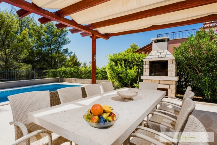 Großzügige Villa im Mittelmeerstil mit 3 SZ, Pool, Dachterrasse mit Meerblick (12) - Ein sonniger Essbereich im Freien mit einem weißen Tisch und Korbstühlen unter einer Pergola. Eine Schale mit Obst steht auf dem Tisch. Im Hintergrund befinden sich ein Steingrill, üppiges Grün und ein Swimmingpool. (45892)