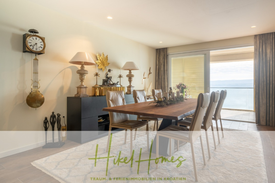MEERBLICK & KUNST Designer Luxus Appartement mit 3 Schlafzimmer – großer Loggia - Modernes Esszimmer mit einem Holztisch, acht Stühlen, dekorativen Lampen, einer Uhr und Blick aufs Meer durch große Schiebetüren. Das Sonnenlicht erhellt den Raum und der Schriftzug "Hikel Homes" erscheint am unteren Rand. (45391)