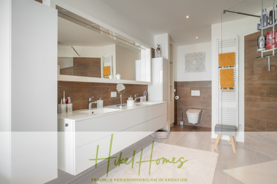 MEERBLICK & KUNST Designer Luxus Appartement mit 3 Schlafzimmer – großer Loggia - Modernes Badezimmer mit weißem Doppelwaschbecken, großem Spiegel, wandmontierter Toilette, Handtuchheizkörper mit orangefarbenen Handtüchern, Glasdusche und braunen Fliesen in Holzoptik. "Hikel Homes"-Logo und Textüberlagerung am unteren Rand. (45373)