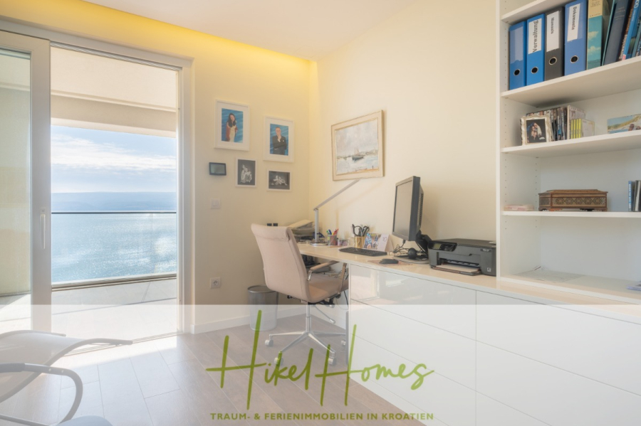 MEERBLICK & KUNST Designer Luxus Appartement mit 3 Schlafzimmer – großer Loggia - Helles Arbeitszimmer mit einem Schreibtisch, einem Computer, Regalen und gerahmten Bildern an der Wand, mit Blick auf ein großes Fenster mit Meerblick. Logo und Text von "Hikel Homes" erscheinen am unteren Rand des Bildes. (45378)