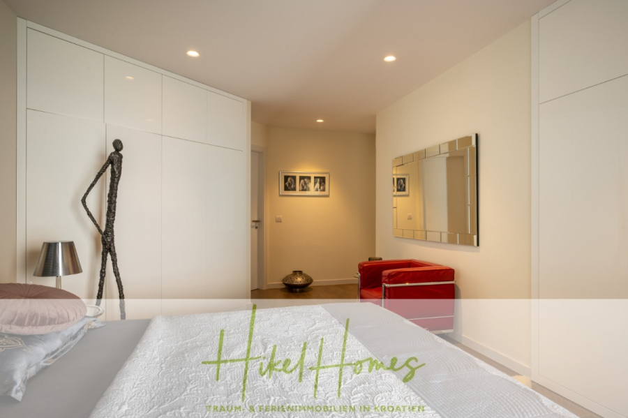 MEERBLICK & KUNST Designer Luxus Appartement mit 3 Schlafzimmer – großer Loggia - Modernes Schlafzimmer mit weißen Wänden und Einbauschränken, einem roten Sessel, einem großen Spiegel, einer Metallskulptur, gerahmten Fotos und einer grauen Tagesdecke. Die stimmungsvolle Beleuchtung und die minimalistische Einrichtung schaffen eine gemütliche, stilvolle Atmosphäre. (45371)