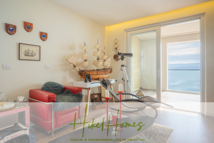 MEERBLICK & KUNST Designer Luxus Appartement mit 3 Schlafzimmer – großer Loggia - Ein helles Zimmer mit einem roten Sessel, einem Schiffsmodell, einem Teleskop mit Blick auf einen Balkon mit Meerblick, einer Wanddekoration im maritimen Stil und einer Glasschiebetür, die Tageslicht hereinlässt. (45374)