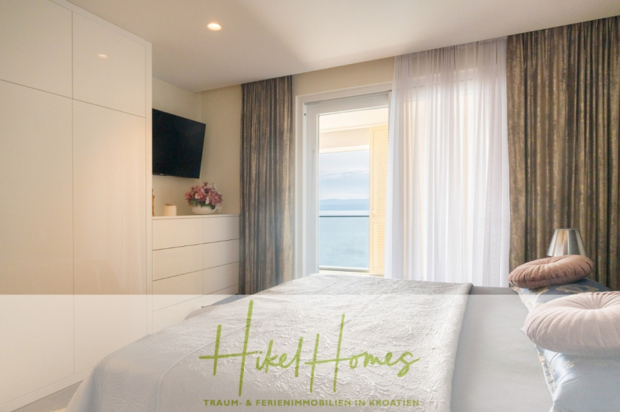 MEERBLICK & KUNST Designer Luxus Appartement mit 3 Schlafzimmer – großer Loggia - Helles, modernes Schlafzimmer mit einem ordentlich gemachten Bett, weißen Möbeln, einem an der Wand montierten Fernseher, Blumen auf einer Kommode, einem großen Fenster mit Vorhängen und einem Blick auf das Meer. Der Text "Hikel Homes" ist am unteren Rand sichtbar. (45370)