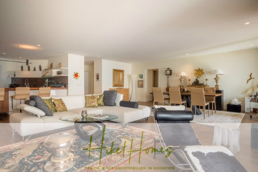 MEERBLICK & KUNST Designer Luxus Appartement mit 3 Schlafzimmer – großer Loggia - Geräumiges, modernes Wohnzimmer mit einem großen weißen Sofa, gemusterten Kissen und einer einzigartigen Chaiselongue. Offene Küche mit Barhockern im Hintergrund. Essbereich mit langem Tisch und eleganten Stühlen. Neutrale Töne und dekorative Akzente prägen das gesamte Raumkonzept. (41084)