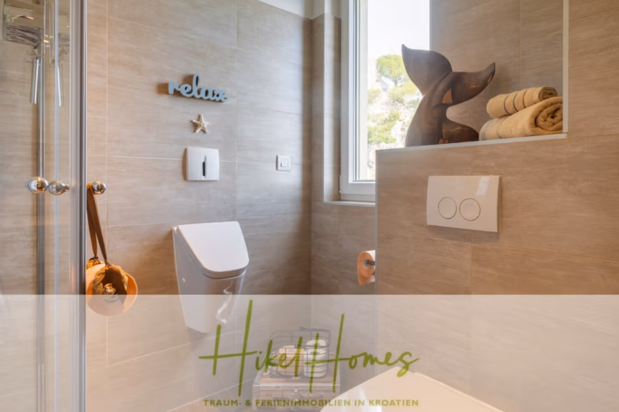MEERBLICK & KUNST Designer Luxus Appartement mit 3 Schlafzimmer – großer Loggia - Modernes Badezimmer mit beige gefliesten Wänden, Wandtoilette und Dusche. An der Wand prangt der Schriftzug „Relax“ mit einem Seestern. Auf einem Regal stehen gerollte Handtücher und eine hölzerne Delfinskulptur. (41078)