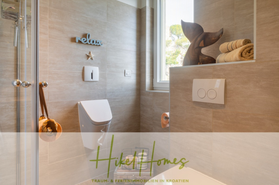 MEERBLICK & KUNST Designer Luxus Appartement mit 3 Schlafzimmer – großer Loggia - Modernes Badezimmer mit beigen Fliesen, einer Dusche, dekorativen Gegenständen wie einem hölzernen Delfin, gerollten Handtüchern, einer an der Wand befestigten Skulptur und dem Wort "Relax" an der Wand über der Toilette. Ein Fenster lässt natürliches Licht herein. (45381)