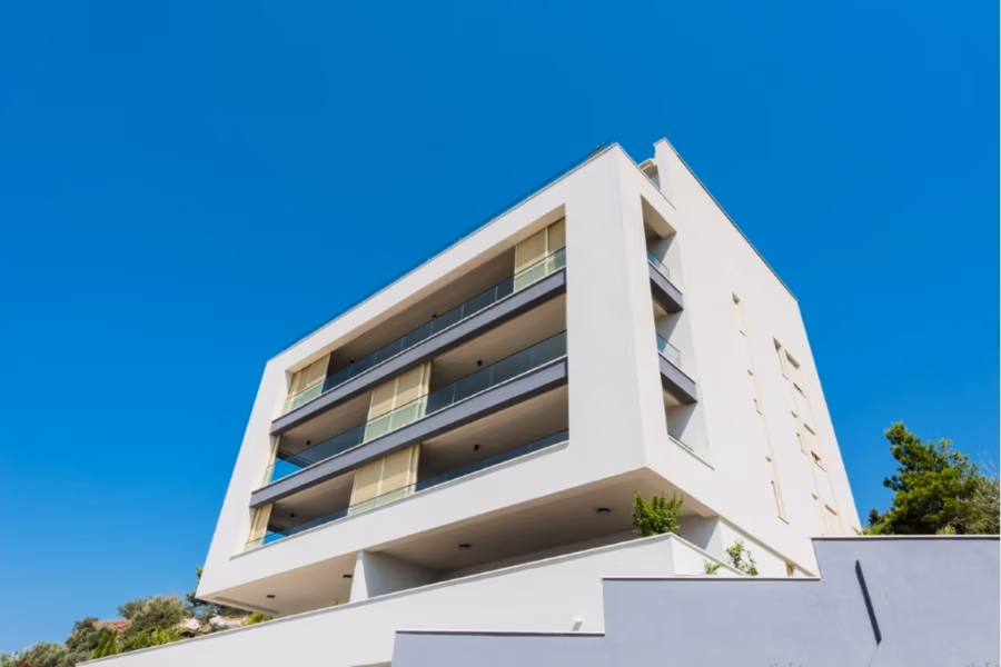 MEERBLICK & KUNST Designer Luxus Appartement mit 3 Schlafzimmer – großer Loggia - Modernes weißes Mehrfamilienhaus mit großen Balkonen und Glasgeländern vor einem strahlend blauen Himmel. Das Gebäude wirkt erhaben und minimalistisch. An den Rändern ist spärliches Grün sichtbar. (41091)
