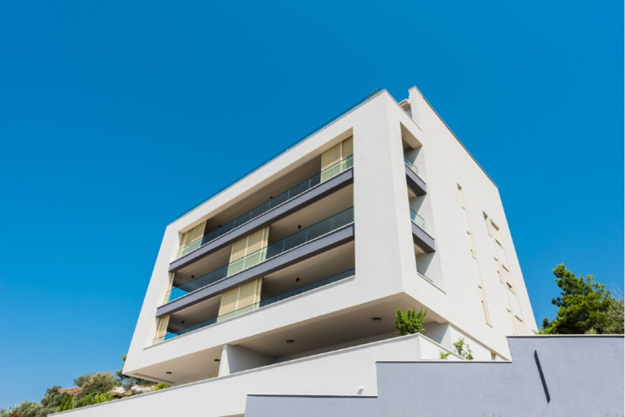 MEERBLICK & KUNST Designer Luxus Appartement mit 3 Schlafzimmer – großer Loggia - Ein modernes weißes Mehrfamilienhaus mit Glasbalkonen, gesehen aus einem niedrigen Winkel vor einem klaren blauen Himmel. Im Vordergrund sind einige Grünpflanzen und eine graue Mauer zu sehen. (45398)