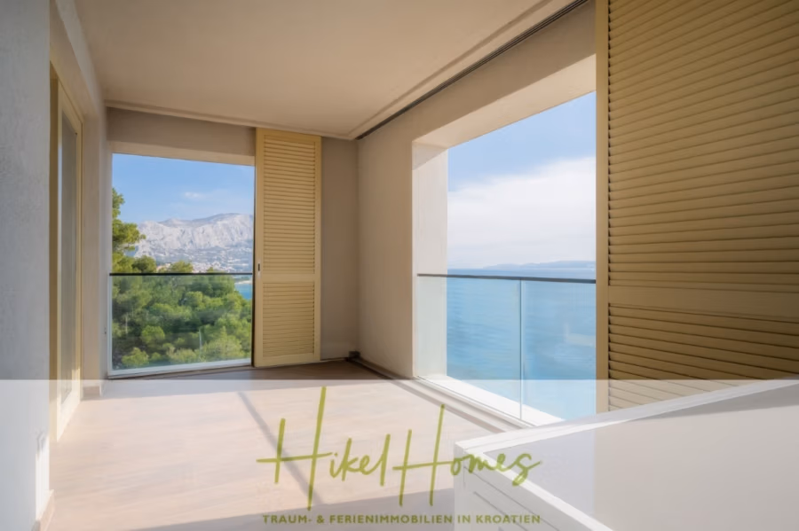 MEERBLICK & KUNST Designer Luxus Appartement mit 3 Schlafzimmer – großer Loggia - Ein Zimmer mit offenen Schiebetüren und Blick auf Meer und Berge. Der Raum ist mit hellem Holzboden und beigefarbenen Wänden ausgestattet. Der Balkon hat ein Glasgeländer, von dem aus Bäume zu sehen sind. Unten steht „Hikel Homes – Traum- & Ferienimmobilien in Kroatien“. (41077)