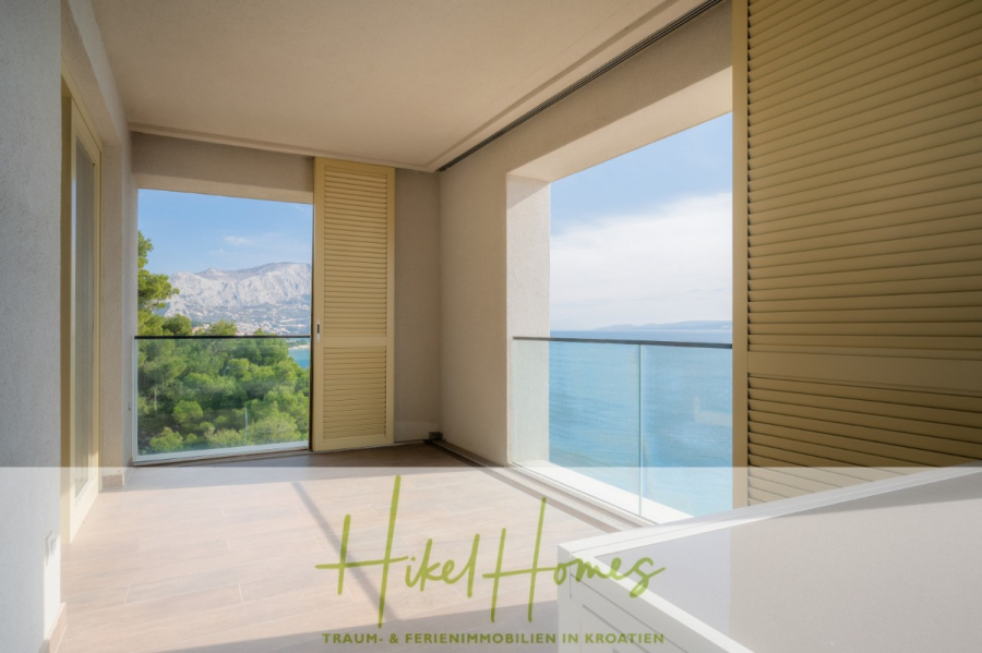 MEERBLICK & KUNST Designer Luxus Appartement mit 3 Schlafzimmer – großer Loggia - Ein heller, moderner Balkon mit hölzernen Fensterläden gibt den Blick auf das Meer, die fernen Berge und das üppige Grün frei. Glasgeländer verschönern die malerische Außenlandschaft. Der Schriftzug "Hikel Homes" erscheint am unteren Rand des Bildes. (45379)