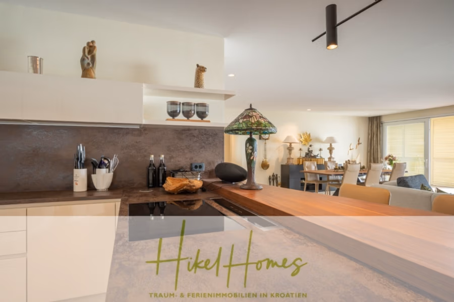 MEERBLICK & KUNST Designer Luxus Appartement mit 3 Schlafzimmer – großer Loggia - Moderne Küche mit glatten Arbeitsflächen und einer Lampe über einem hölzernen Esstisch. Der offene Grundriss führt zum Wohnbereich mit Dekoration und Tageslicht. Unten steht „Hikel Homes – Traum & Ferienimmobilien in Kroatien“. (41090)