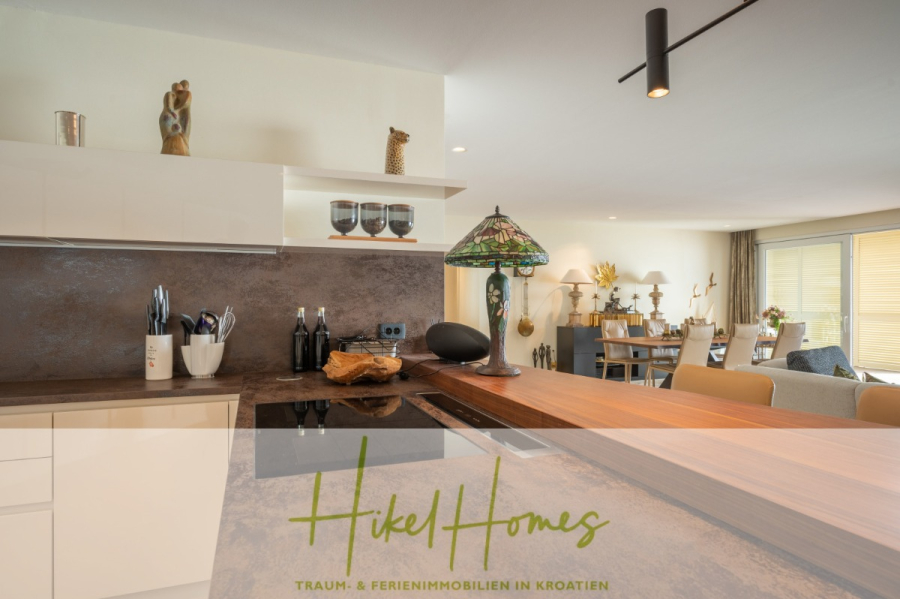 MEERBLICK & KUNST Designer Luxus Appartement mit 3 Schlafzimmer – großer Loggia - Moderne Küche mit brauner Arbeitsplatte, Utensilien, Weinflaschen und Dekorationsartikeln. Der Raum öffnet sich zu einem hellen, stilvollen Wohn- und Essbereich. "Hikel Homes: Traum- & Ferienimmobilien in Kroatien" ist am unteren Rand eingeblendet. (45397)