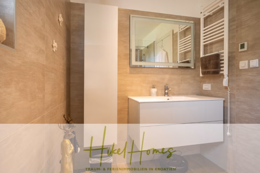 MEERBLICK & KUNST Designer Luxus Appartement mit 3 Schlafzimmer – großer Loggia - Modernes Badezimmer mit beige gefliesten Wänden, einem weißen Waschtisch mit Spiegel und einem schlichten, minimalistischen Design. Ein Handtuch hängt an einem beheizten Ständer, und eine kleine dekorative Statue steht auf dem Boden. Unten ist der grüne Text „Hikel Homes“ eingelegt. (41079)