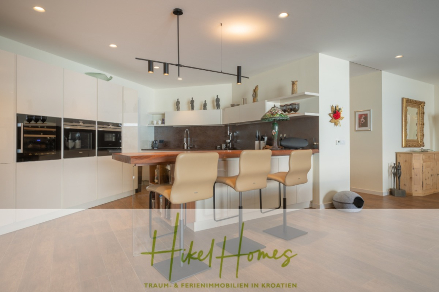 MEERBLICK & KUNST Designer Luxus Appartement mit 3 Schlafzimmer – großer Loggia - Moderne Küche mit weißen Schränken, Einbaugeräten, einer großen Holzinsel mit vier beigen Barhockern, dekorativen Regalen und sanfter Beleuchtung. Der Schriftzug "Hikel Homes" ist am unteren Rand des Bildes eingeblendet. (45394)