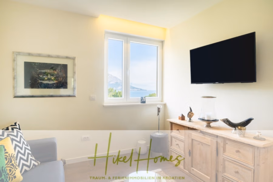 MEERBLICK & KUNST Designer Luxus Appartement mit 3 Schlafzimmer – großer Loggia - Helles Wohnzimmer mit wandmontiertem Fernseher und einem mit Dekoartikeln geschmückten Holz-Sideboard. Ein großes Fenster bietet Blick auf Berge und Meer. Daneben steht ein graues Sofa mit gelb gemusterten Kissen. Unten steht der Schriftzug „Hikel Homes“. (41075)