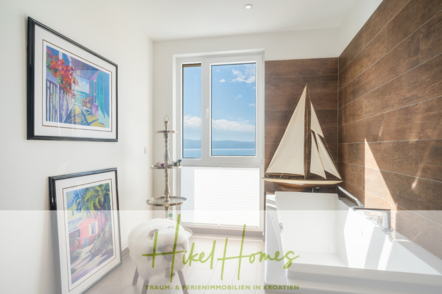 MEERBLICK & KUNST Designer Luxus Appartement mit 3 Schlafzimmer – großer Loggia - Helles Badezimmer mit einem großen Fenster mit Meerblick, einer weißen Badewanne, einem dekorativen Modellsegelboot, zwei bunten gerahmten Bildern an der Wand und einer Holzverkleidung hinter der Badewanne. (45369)