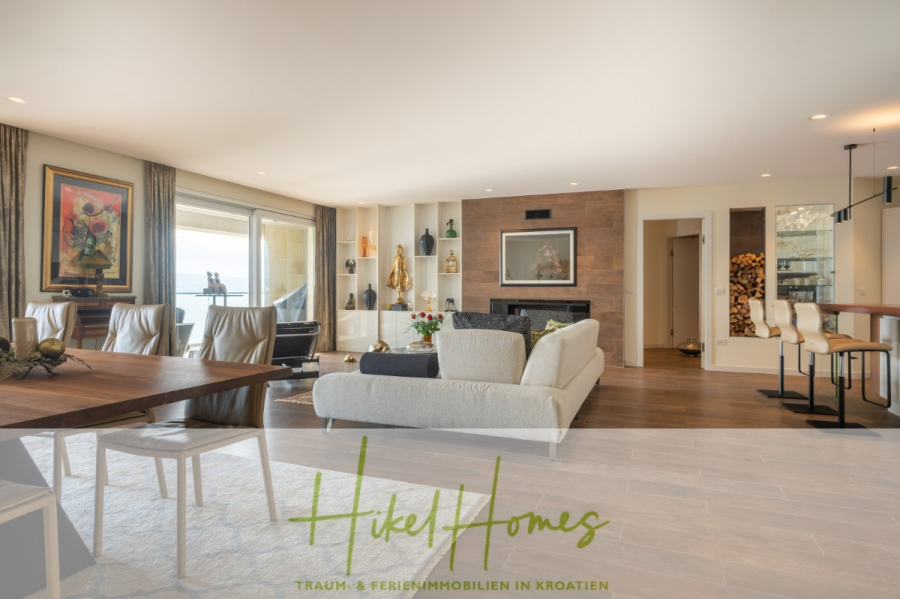 MEERBLICK & KUNST Designer Luxus Appartement mit 3 Schlafzimmer – großer Loggia - Geräumiger moderner Wohn- und Essbereich mit neutralen Farbtönen, großen Fenstern, Kunstwerken, offener Küche mit Barhockern und dekorativen Regalen. Das Logo "Hikel Homes" und ein Text-Overlay erscheinen am unteren Rand. (45390)