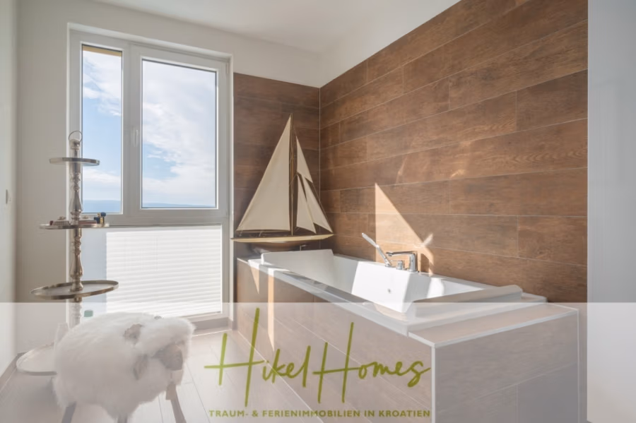 MEERBLICK & KUNST Designer Luxus Appartement mit 3 Schlafzimmer – großer Loggia - Das helle Badezimmer mit Holzakzentwand verfügt über eine weiße Badewanne, ein Segelbootmodell und einen flauschigen Hocker. Sonnenlicht fällt durch ein großes Fenster und erhellt den Raum. Der Text lautet: „Hikel Homes – Traum- & Ferienimmobilien in Kroatien.“. (41074)