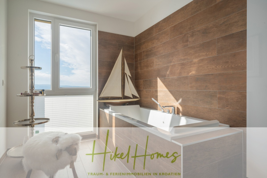 MEERBLICK & KUNST Designer Luxus Appartement mit 3 Schlafzimmer – großer Loggia - Ein modernes Badezimmer mit einem großen Fenster, einer rechteckigen Badewanne, hölzernen Wandfliesen, einem dekorativen Segelboot, einem verchromten Handtuchständer und einem Hocker mit einem Sitz aus Fell. Der Schriftzug "Hikel Homes" ist am unteren Rand eingeblendet. (45375)